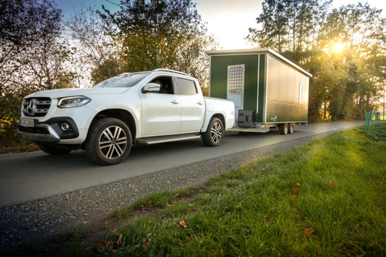 Ein weißer Pickup zieht einen mobilen Hühnerstall in grüner Farbe auf einem Landweg, umgeben von Bäumen bei Sonnenuntergang.