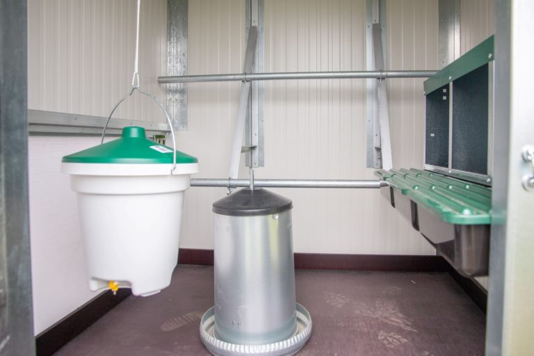 Der Innenraum eines mobilen Hühnerstalls mit einer hängenden Futterstation, einem Wassertank und einer Sitzstange.