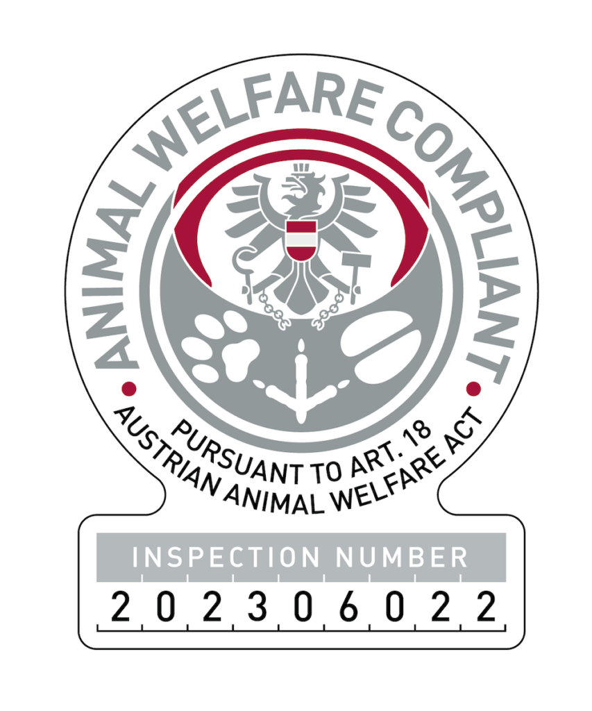 Das Bild zeigt ein Emblem zur Kennzeichnung der Einhaltung des österreichischen Tierschutzgesetzes, das das Wappen mit den Aufschriften „ANIMAL WELFARE COMPLIANT“ und einer Prüfnummer enthält.