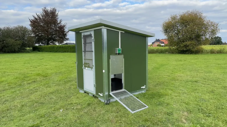 Ein grüner Mobilstall steht auf einer Wiese, umgeben von Bäumen und einer Hecke, mit einer Rampe an der Vorderseite.