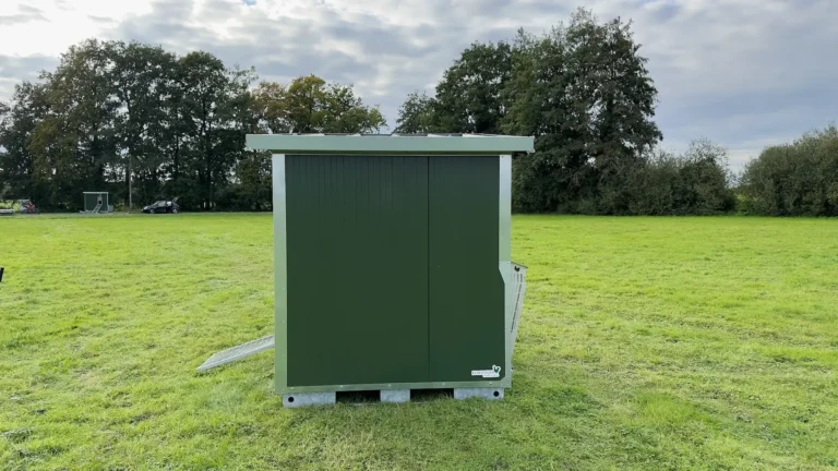 Ein grüner Mobilstall steht auf einer Wiese mit Bäumen im Hintergrund.