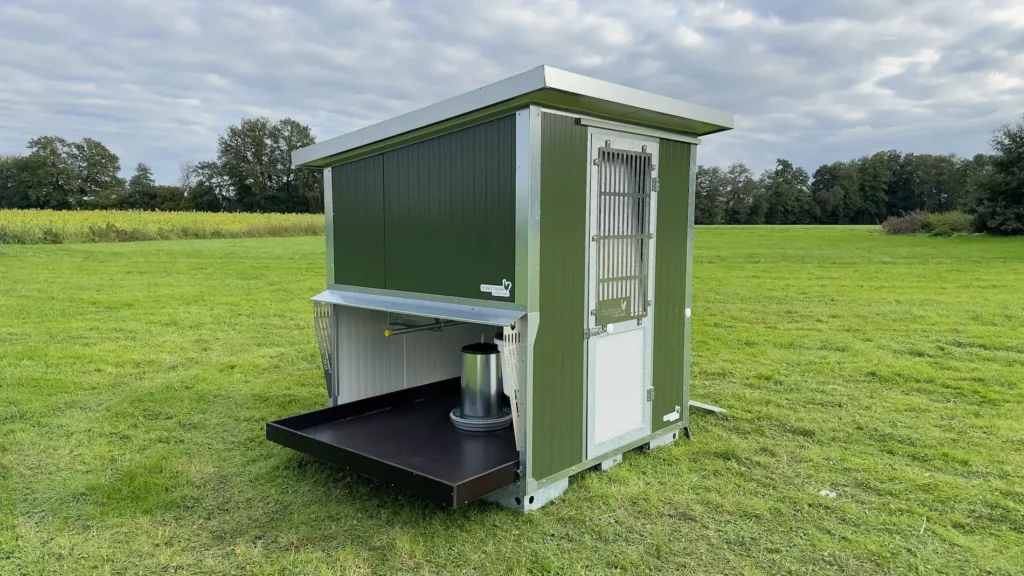 Ein mobiler Stall in grüner und silberner Farbe steht auf einer Wiese unter einem bewölkten Himmel.
