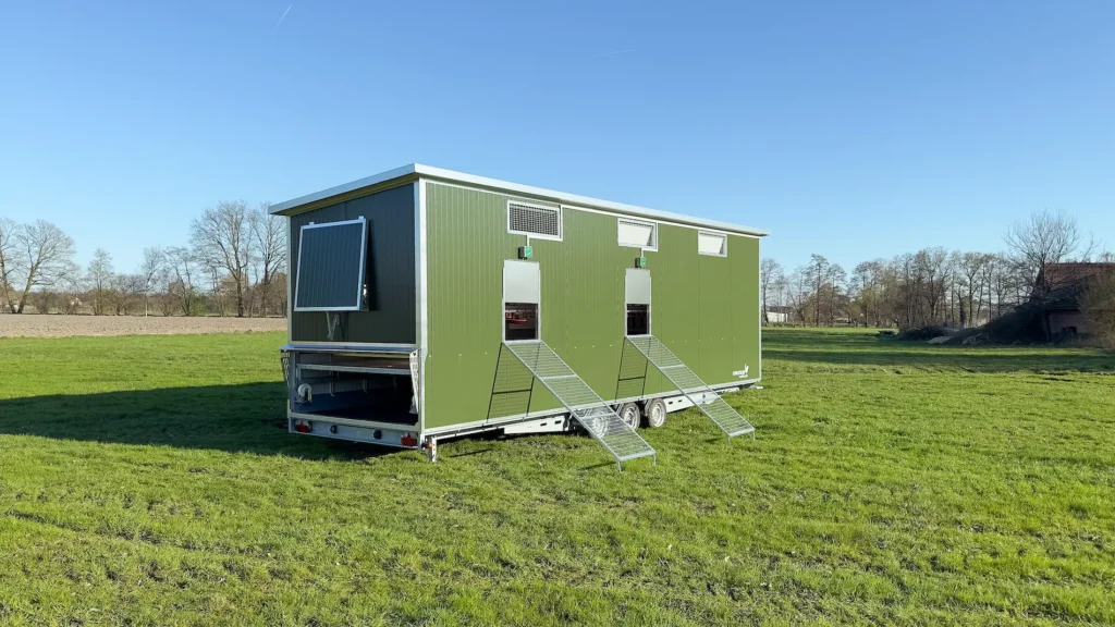 Ein mobiler Hühnerstall in grün steht auf einer Wiese unter blauem Himmel, mit Rampen an den Eingängen für die Hühner.